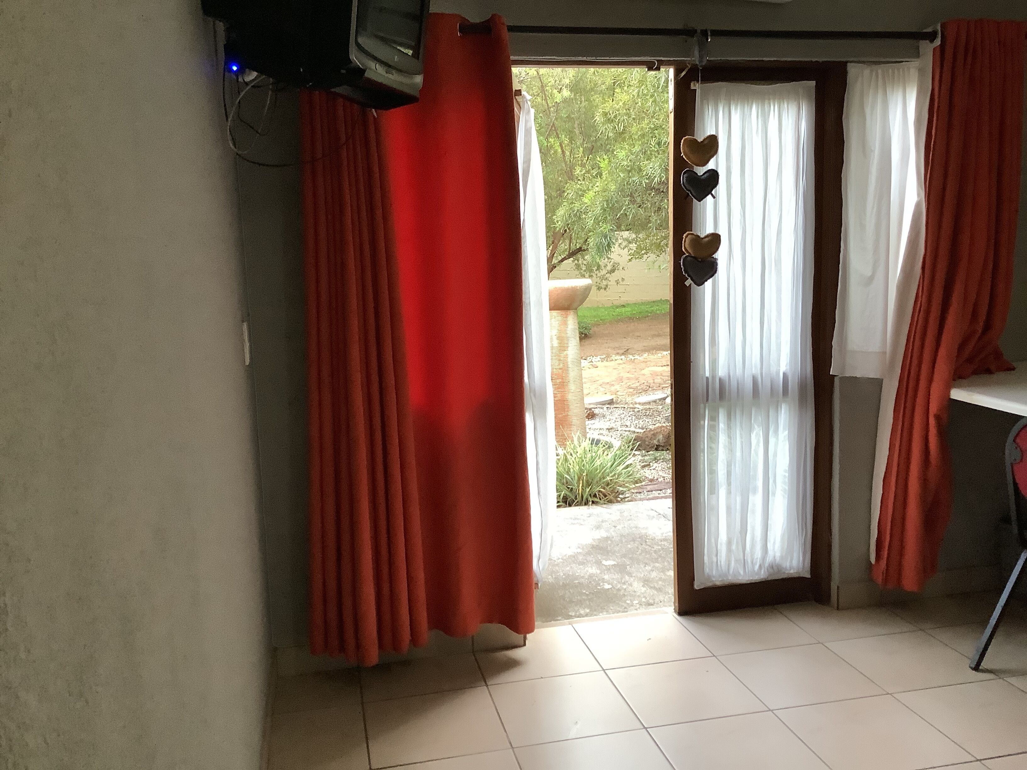 A room with a sliding glass door leading to an outdoor area, red curtains, a white tiled floor, and a small table with a chair.
