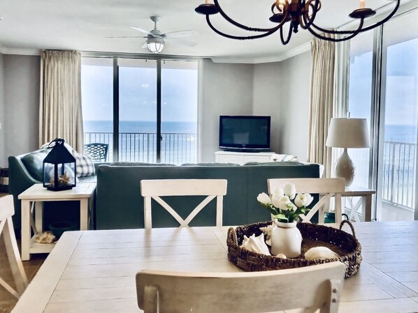 Una habitación de hotel moderna con balcón, zona de comedor con mesa y sillas, sofá, televisor y candelabro.