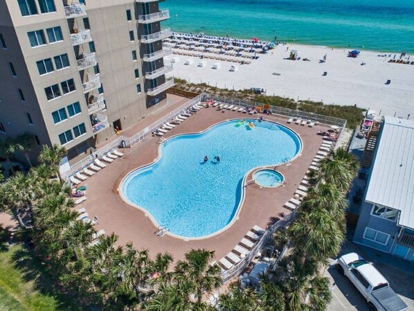 An aerial view of a hotel with a large swimming pool, a beach area, and a parking lot.