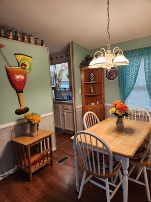 A dining area with a wooden table and white chairs, a kitchen in the background, and a decorative wall with jars and a clock.