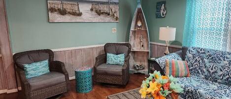A living room with two dark wicker chairs, a patterned sofa, a wooden coffee table with a vase of flowers, and a wall-mounted shelf with decorative items.