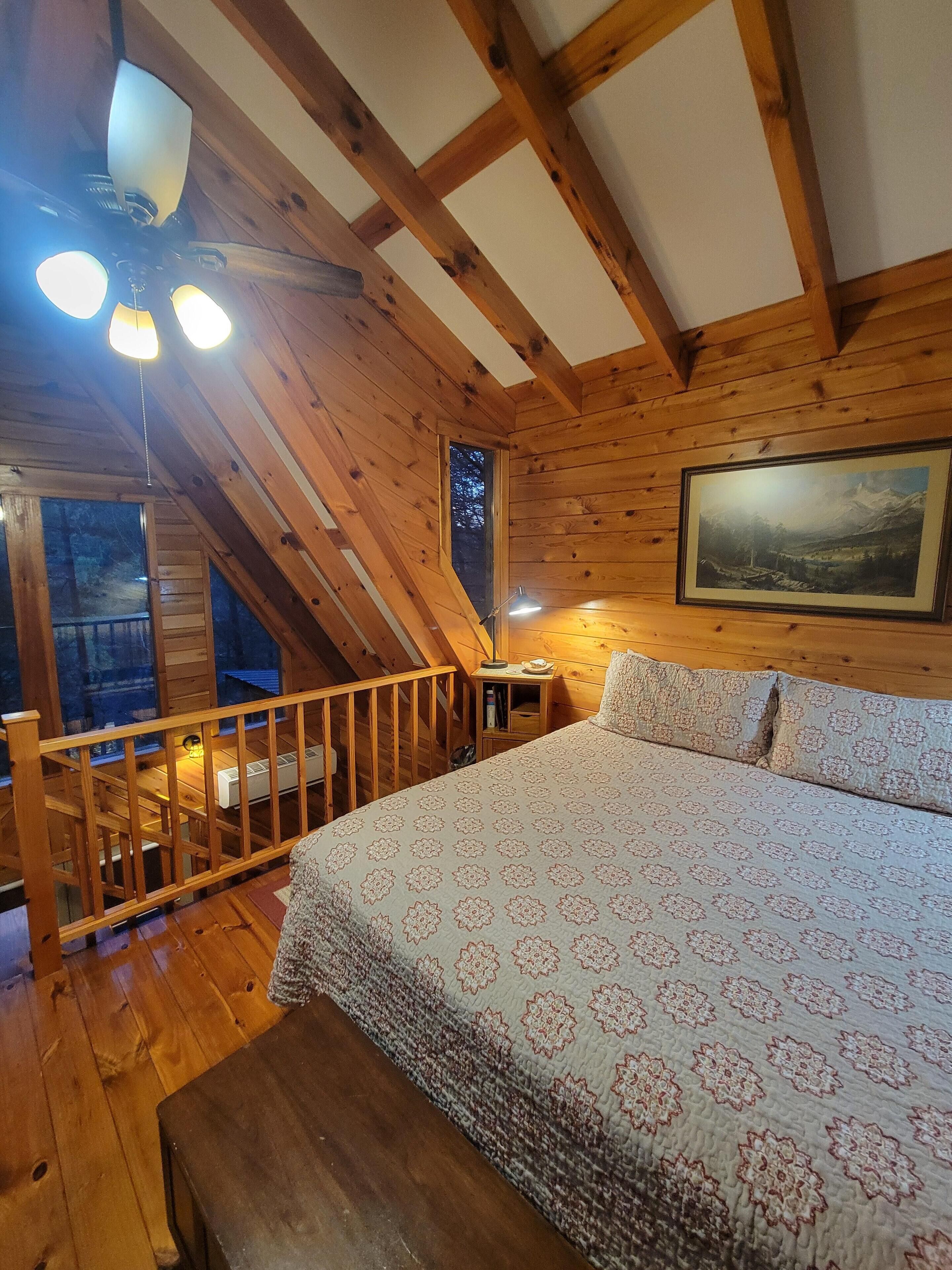 Un dormitorio acogedor en el ático con techo de madera, una cama grande con ropa de cama floral, un cuadro en la pared y una ventana con vistas a árboles.