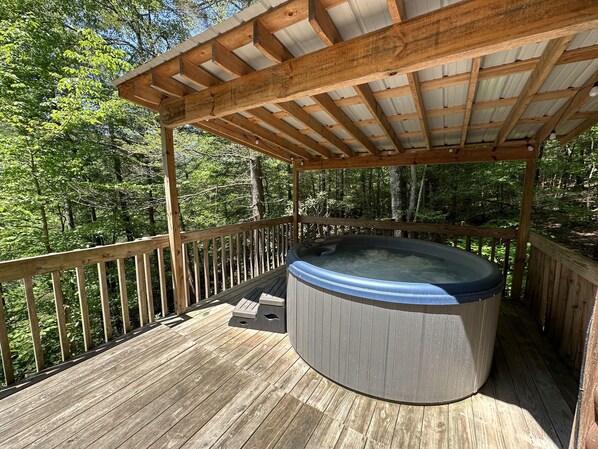 Un jacuzzi en una terraza de madera con una pérgola encima, rodeado de zona boscosa.