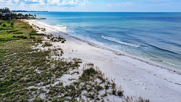 A beach with white sand, clear blue water, and a few buildings in the background.