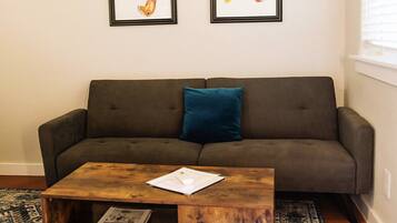 Un salon avec un canapé gris foncé, un coussin bleu, une table basse en bois sur laquelle repose un magazine, et deux œuvres d’art encadrées accrochées au mur.