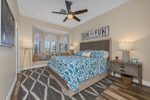 A bedroom with a bed, a ceiling fan, a nightstand, a lamp, a mirror, and a sign that says "SUN AND FUN".
