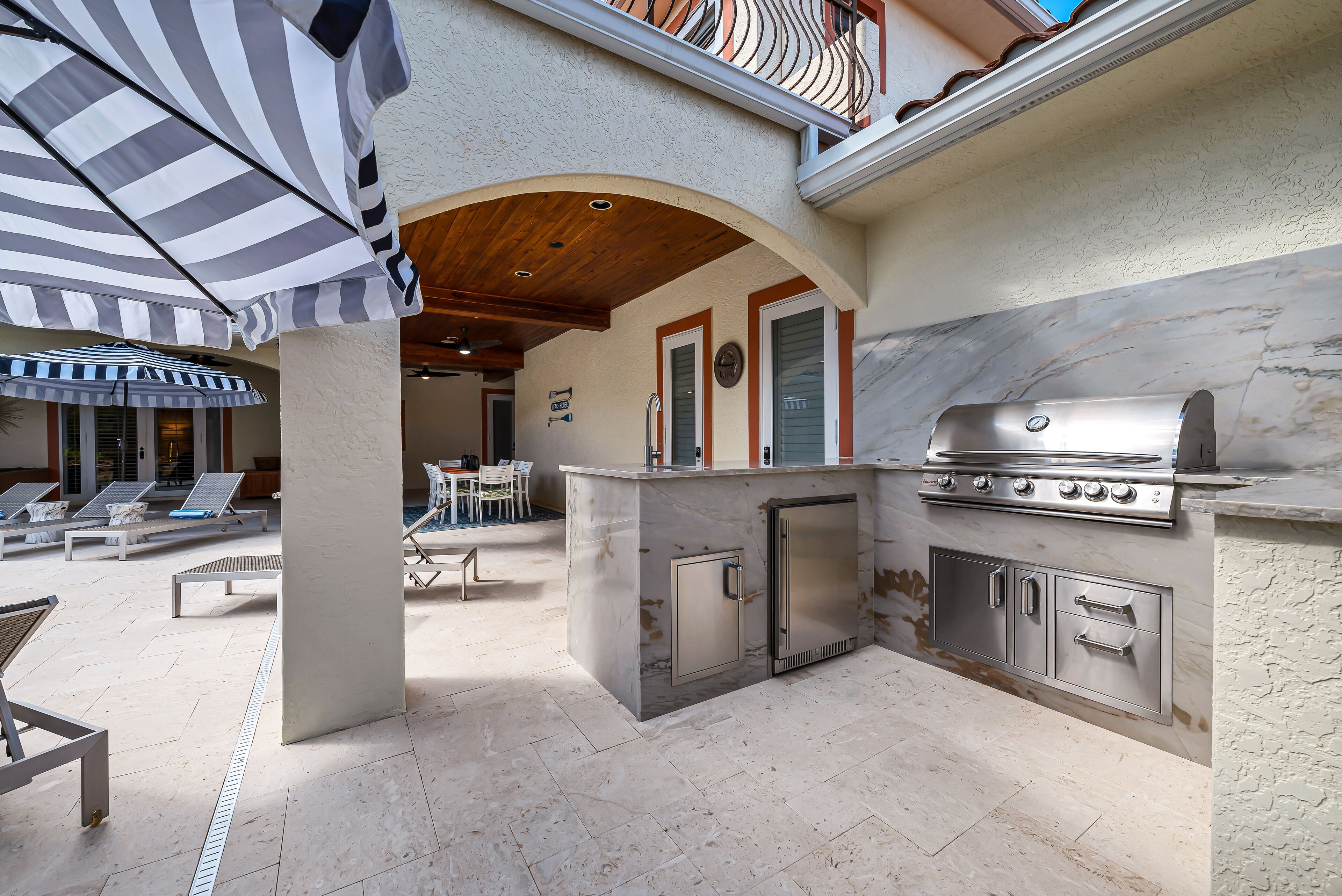 Ein Outdoor-Kochbereich mit Grill, einem Esstisch mit Stühlen und einem gestreiften Sonnenschirm, der Schatten spendet.