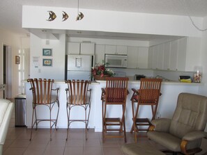 A kitchen with white cabinets, a refrigerator, and a microwave. There are three wooden chairs with cushions and a beige sofa.