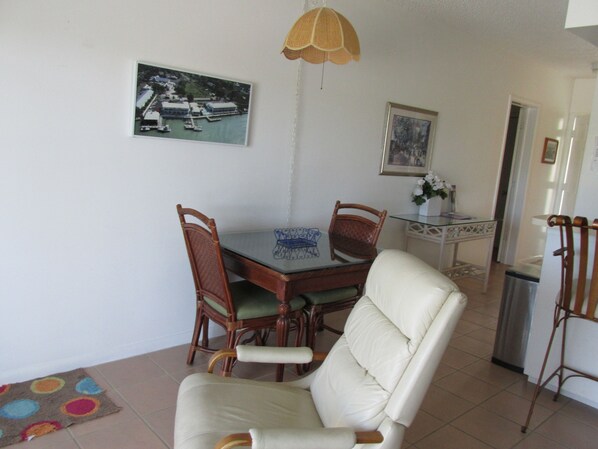 A dining area with a glass table, two wooden chairs, a white cushioned chair, a hanging lamp, and a framed picture on the wall.