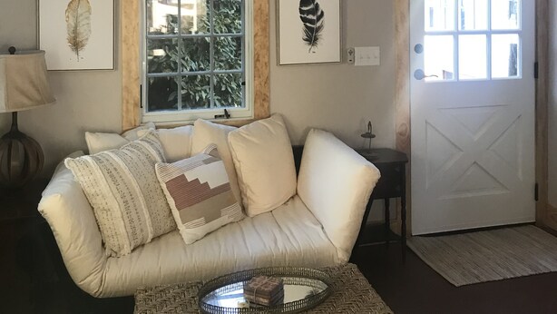 A living room with a beige sofa, a woven coffee table, and a large window with a view of greenery. There are framed pictures on the wall and a door with a window. - Beautiful Asheville Tiny Home Get-a-way (Asheville)