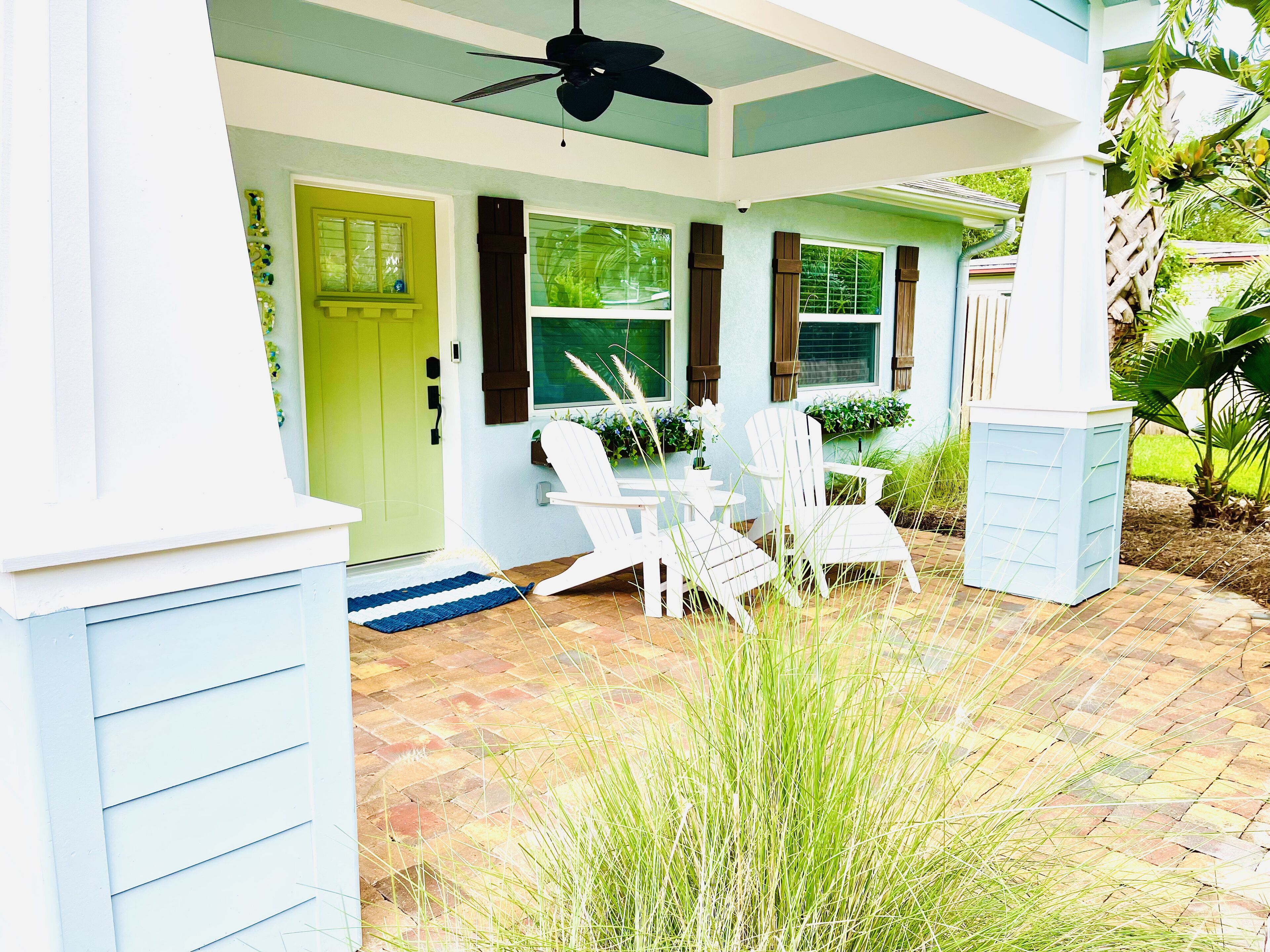 A covered porch with white chairs and a table, a green door, and a ceiling fan.