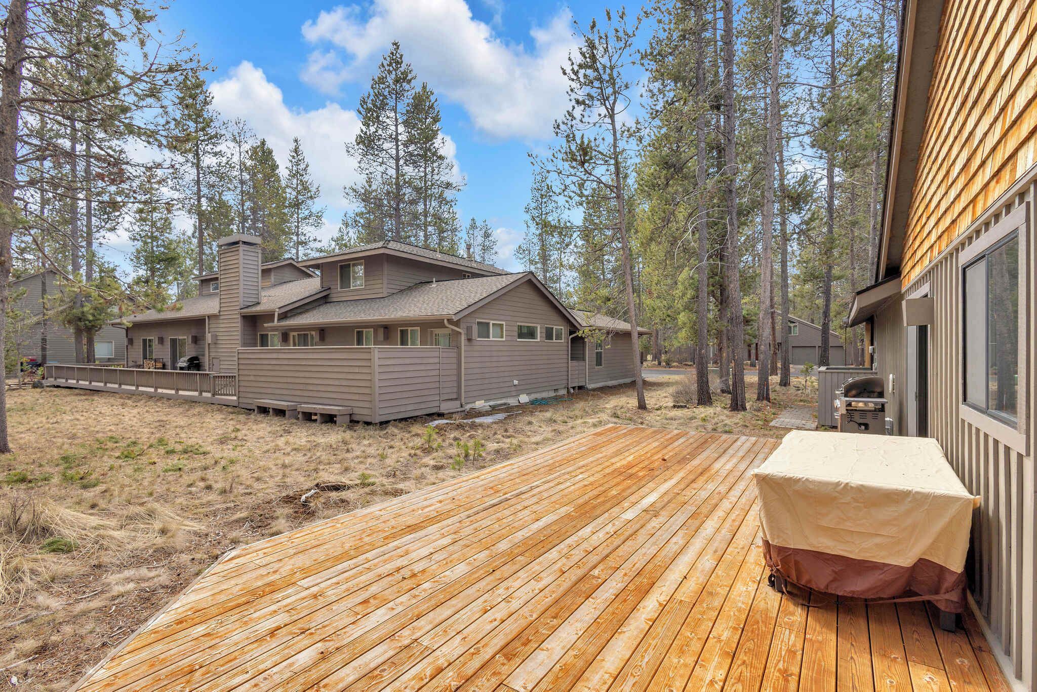 A wooden deck leading to a house with a covered grill area, surrounded by tall pine trees.