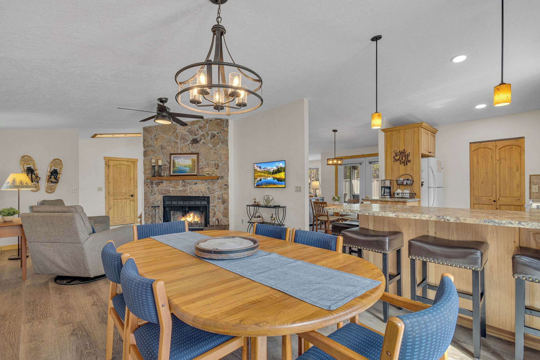 A spacious living room with a fireplace, a dining area with a wooden table and blue chairs, and a kitchen with wooden cabinets and a bar area.