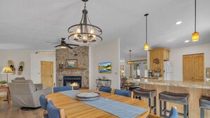 Uma sala de estar ampla com lareira, uma área de jantar com mesa de madeira e cadeiras azuis, e uma cozinha com armários de madeira e um balcão.