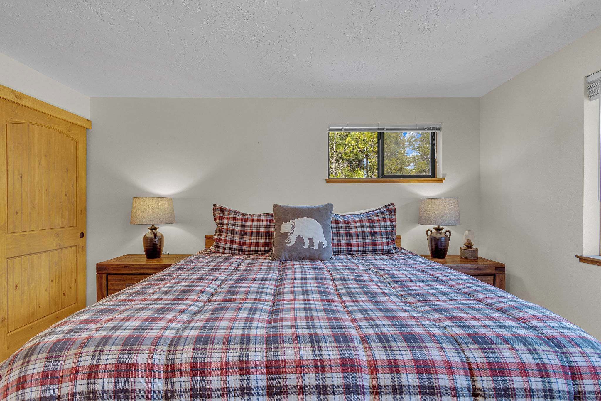 A bedroom with a plaid bedspread, wooden wardrobe, two bedside tables with lamps, and a window with a view of trees.