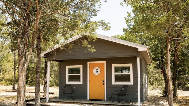 Une petite cabane avec une seule porte et deux fenêtres, entourée d’arbres.