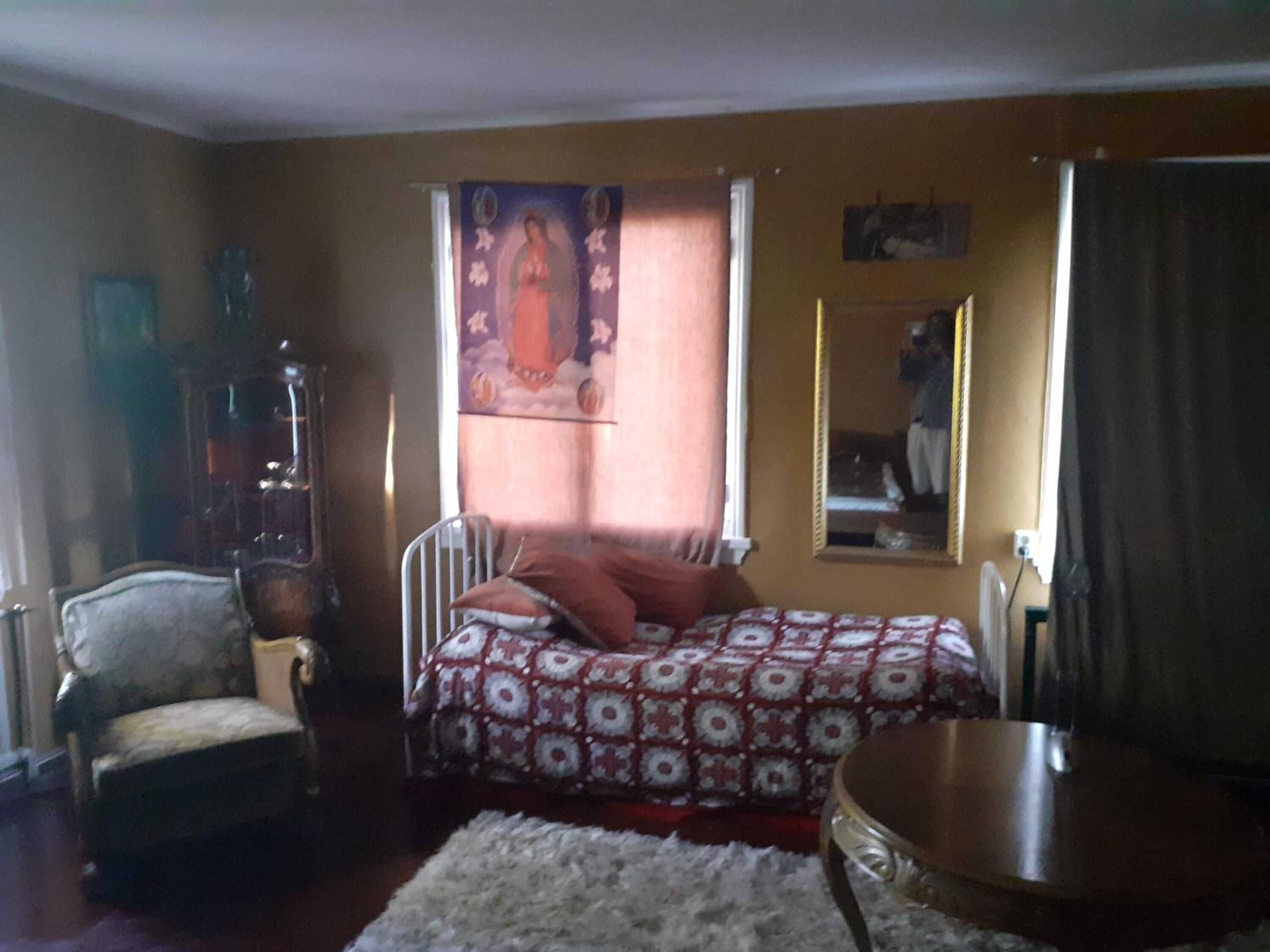 Un dormitorio con cama, una silla, una mesita, un espejo y una ventana con cortinas.