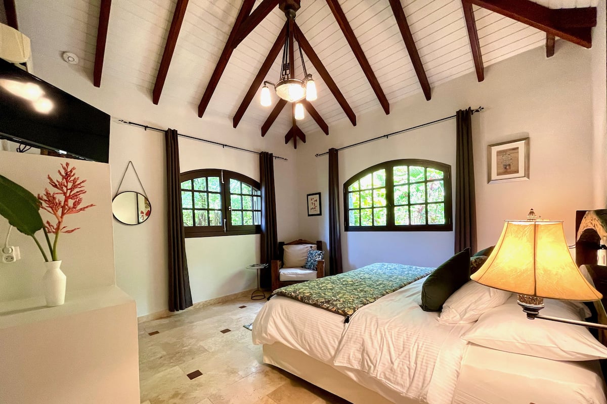 A bedroom with a bed, a bedside lamp, a mirror, a window, and a plant.