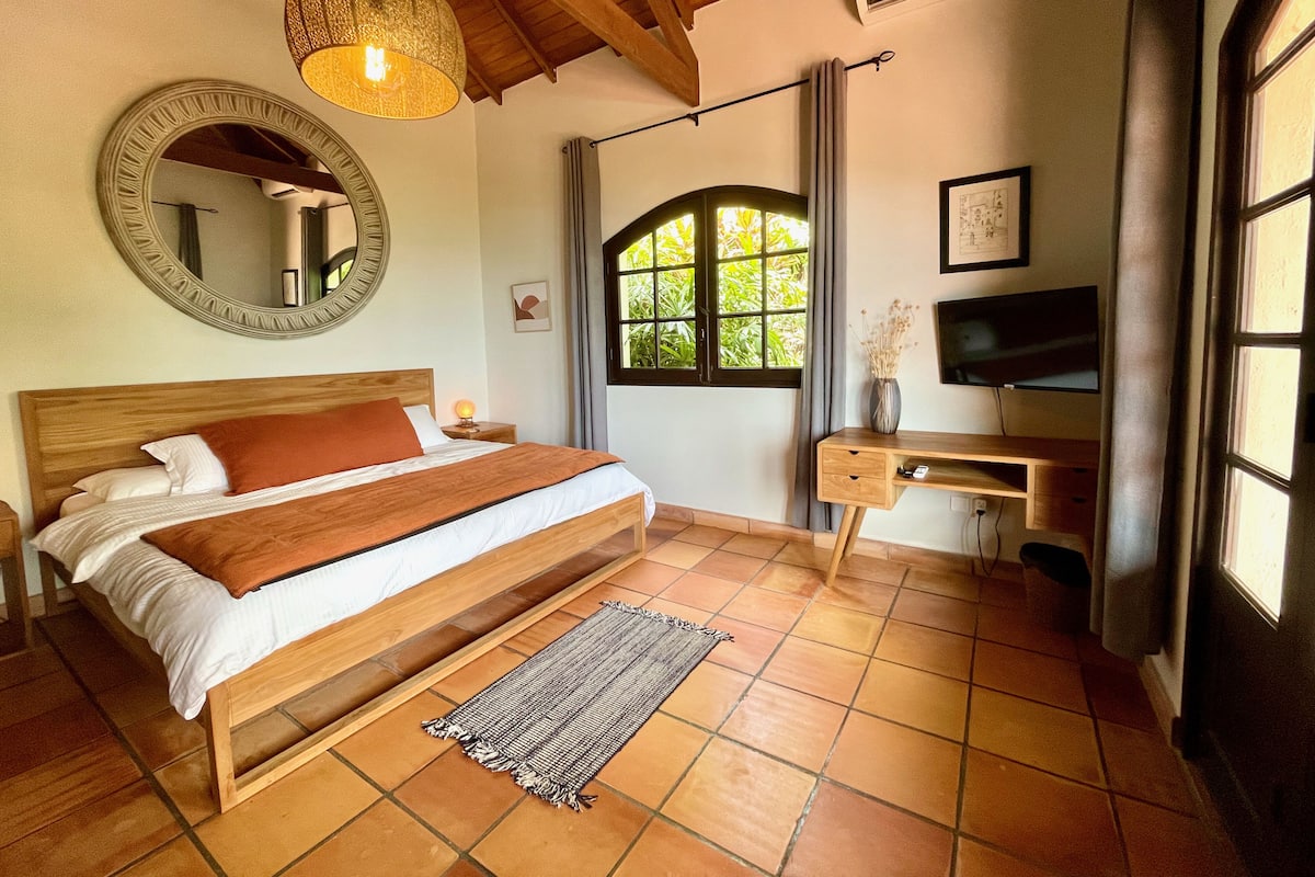 A bedroom with a wooden bed, a desk, a television, a mirror, and a window with curtains.