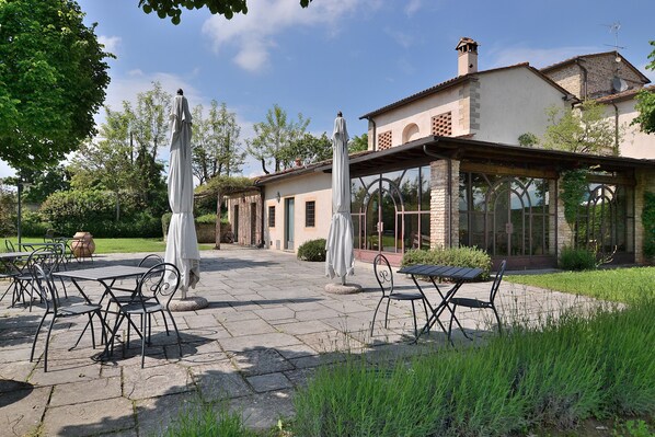 Un patio exterior con suelo de piedra, sillas de metal y sombrillas, rodeado de vegetación y un edificio de grandes ventanales y fachada de ladrillo.