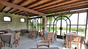 Un espace de restauration en plein air avec des murs en pierre, des tables et des chaises en bois, et de grandes fenêtres offrant une vue sur la verdure.