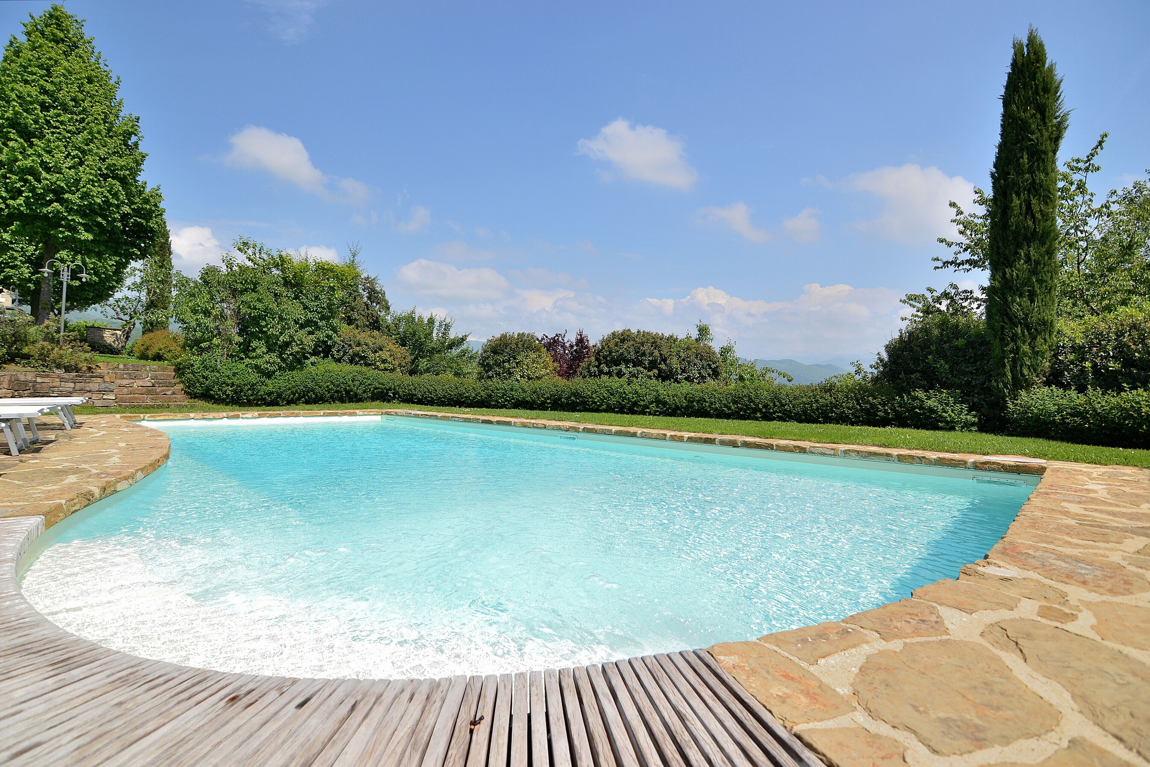 A swimming pool with a wooden deck, surrounded by stone walls and lush greenery.
