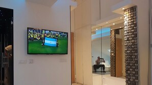 Eine moderne Hotellobby mit einem Flachbildfernseher, der ein Fußballspiel zeigt, einer Glastrennwand und einem Spiegel, der eine Person beim Fotografieren reflektiert.