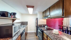 Cocina compacta equipada con microondas, lavavajillas, refrigerador y estufa. Cuenta con gabinetes de madera, encimeras de granito y una pared de ladrillo rojo.