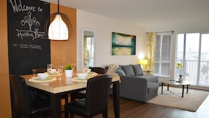 En moderne stue med spiseplads, med et negerbord med en velkomstbesked, et stort vindue med persienner, en grå sofa og et lille bord med en lampe.