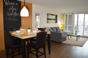 Ein modernes Wohnzimmer mit Essbereich, inklusive einer Tafel mit einer Willkommensbotschaft, einem großen Fenster mit Jalousien, einem grauen Sofa und einem kleinen Tisch mit Lampe.