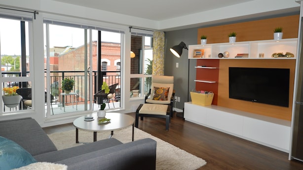 En moderne stue med stort fjernsyn, sofa, lænestol og balkon med udsigt til bygninger.