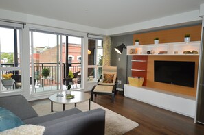A modern living room with a large TV, a sofa, a chair, and a balcony with a view of buildings.