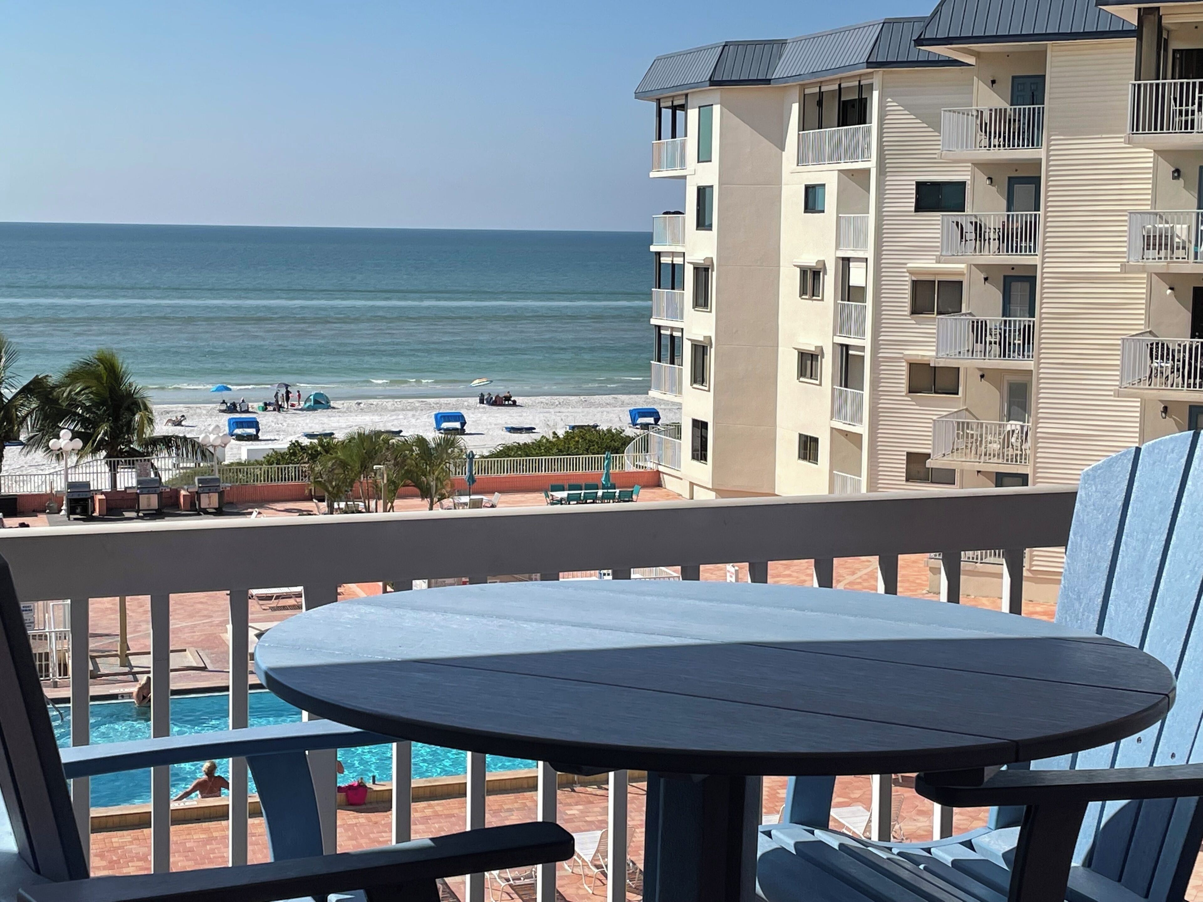 A balcony with a view of the beach and ocean, a pool area, and a building with multiple balconies.