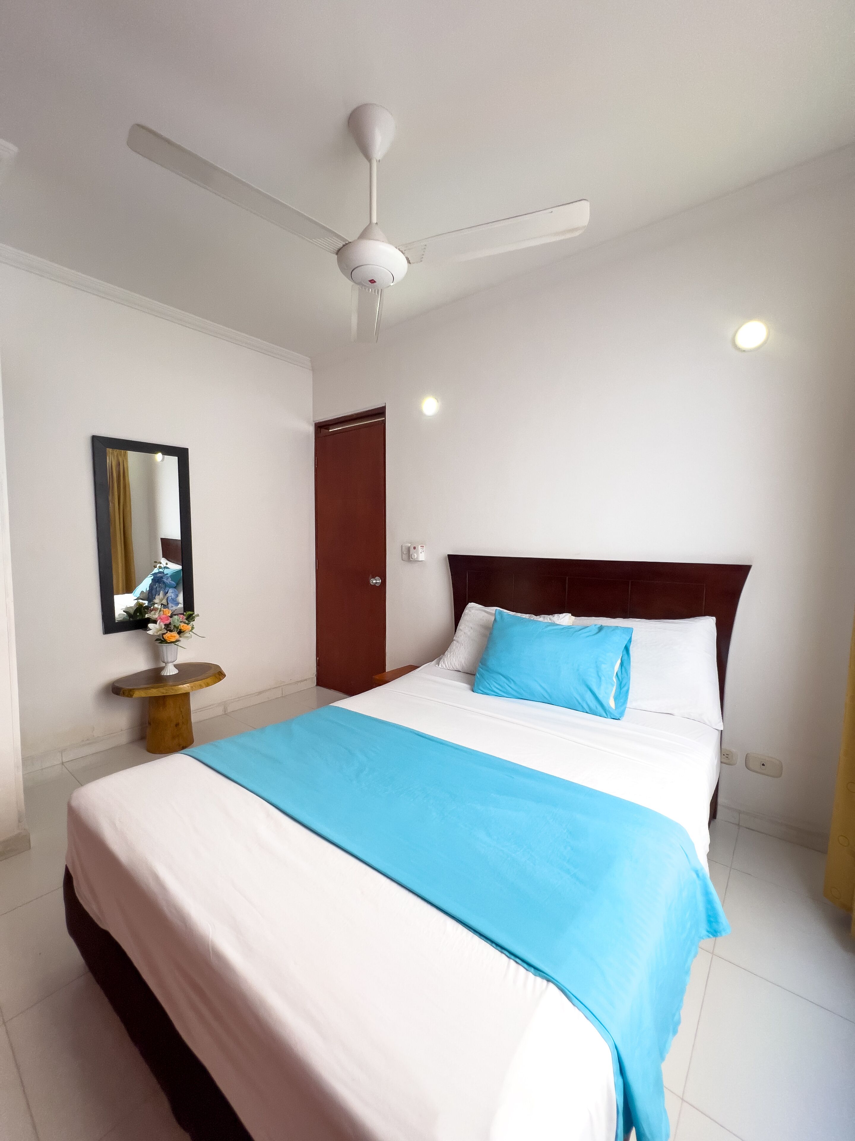 A hotel room with a bed, a ceiling fan, a mirror, a small table with a vase of flowers, and a door.