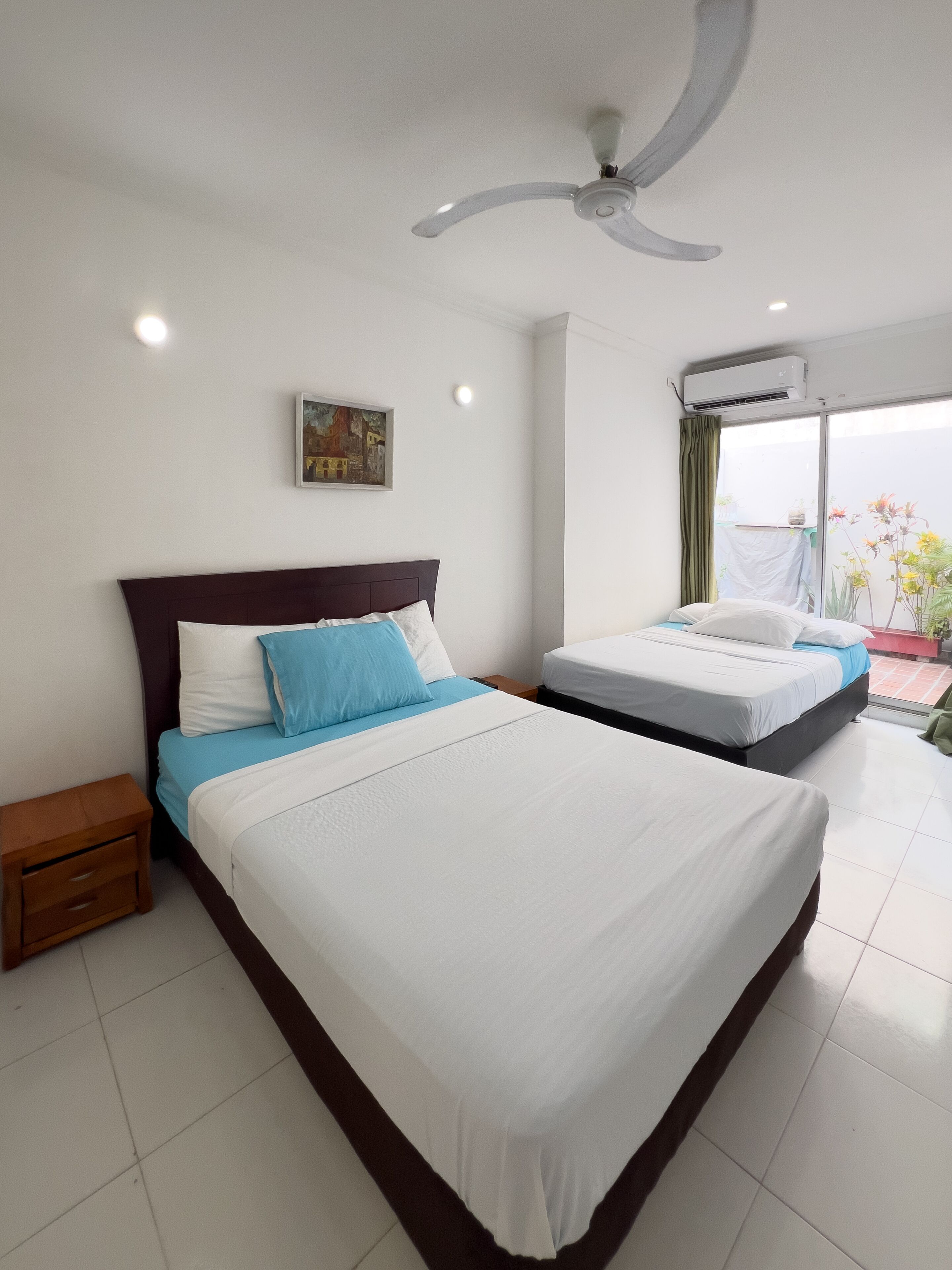 Habitación de hotel con dos camas, ventilador de techo, unidad de aire acondicionado y vista al exterior.