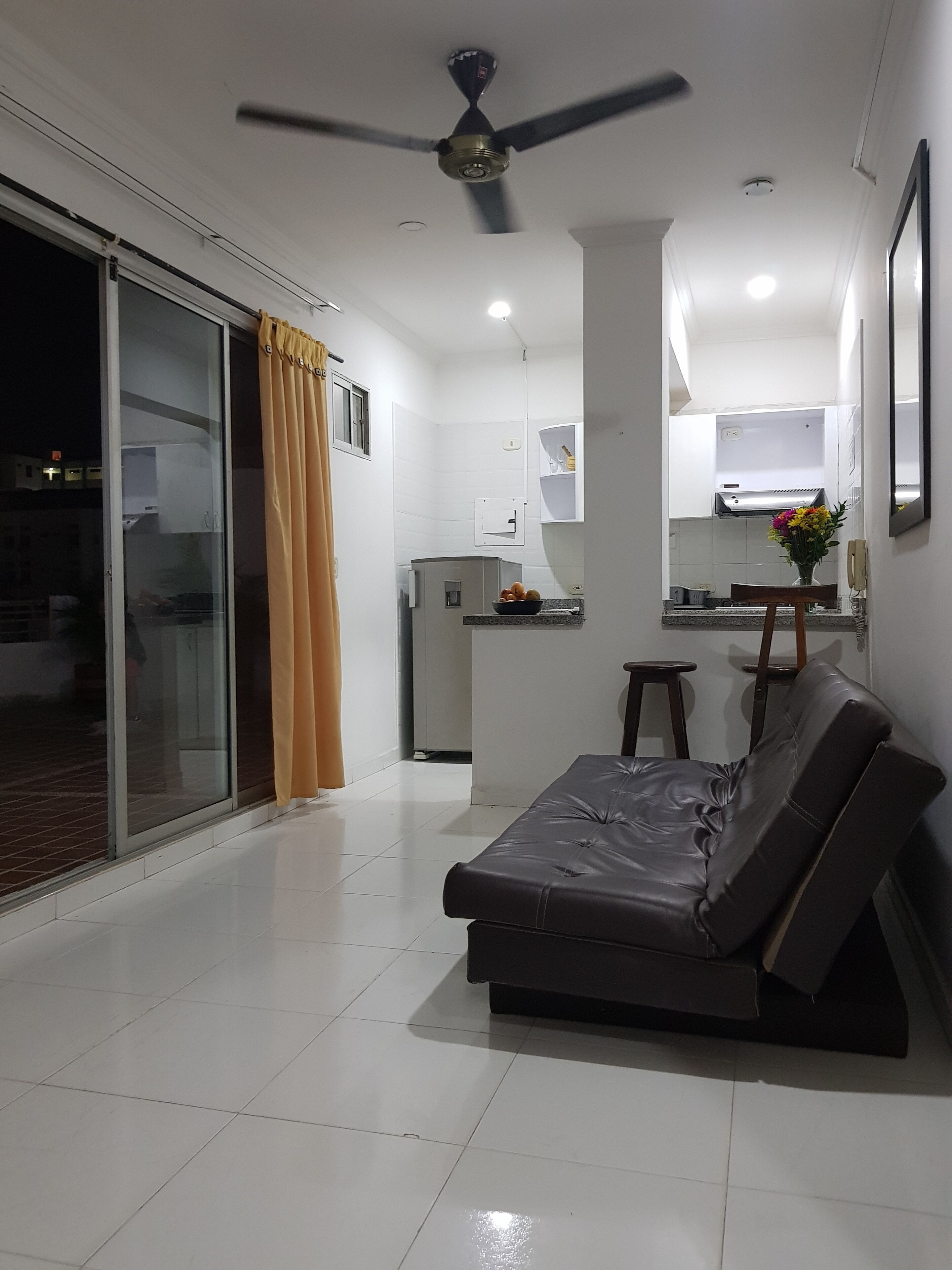 Una sala de estar moderna con un sofá, un ventilador de techo y una cocina al fondo.