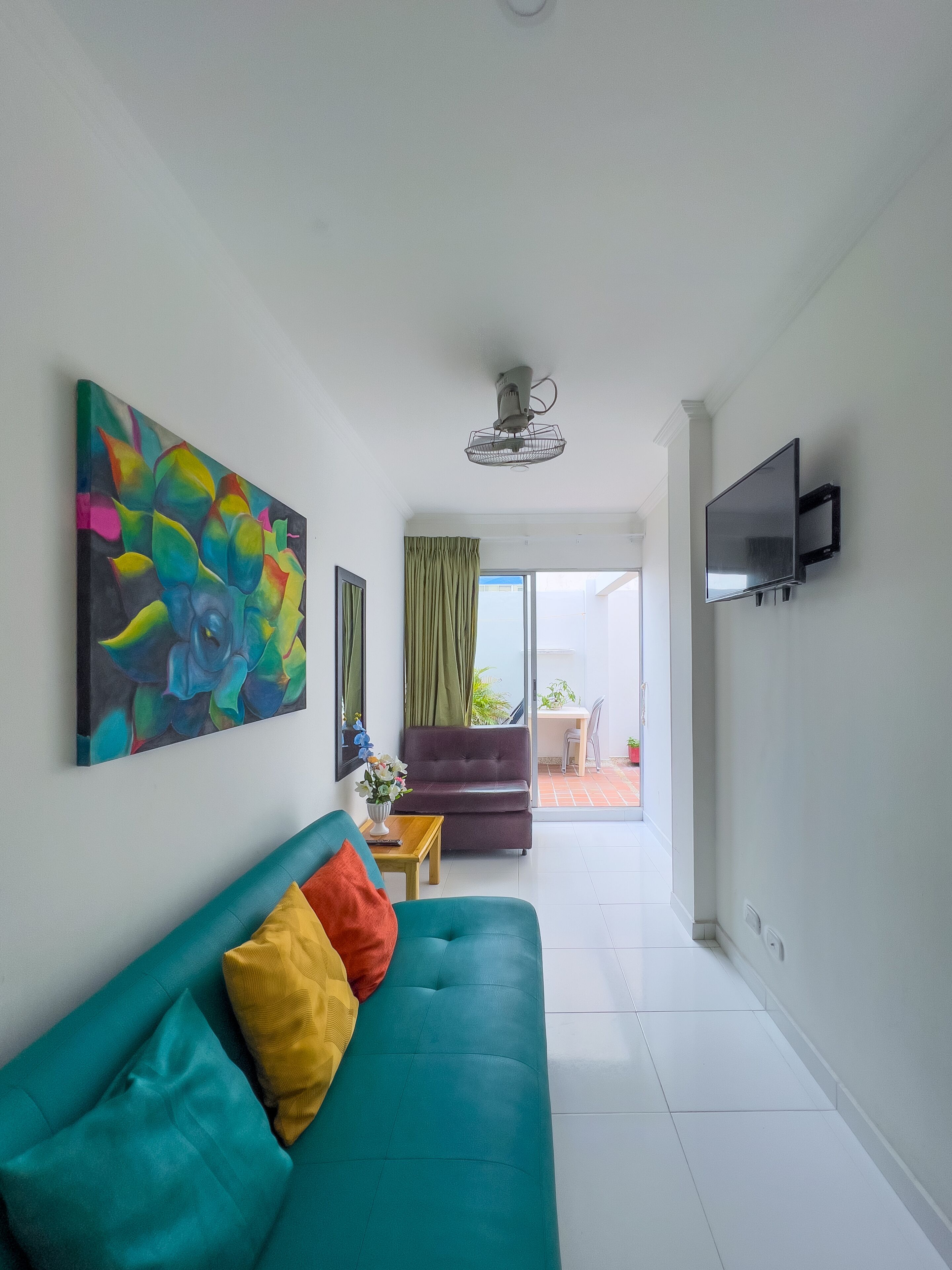 A modern living room with a teal sofa, a colorful abstract painting, a ceiling fan, and a TV mounted on the wall.