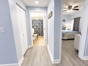 A hallway leading to a bathroom with a toilet and shower, a bedroom with a bed and a ceiling fan, and a sign that reads 'BEACH HOUSE'. - 2128 Pool & Beach views,  w/ two parking passes, spectacular lagoon-style pool (Panama City Beach)