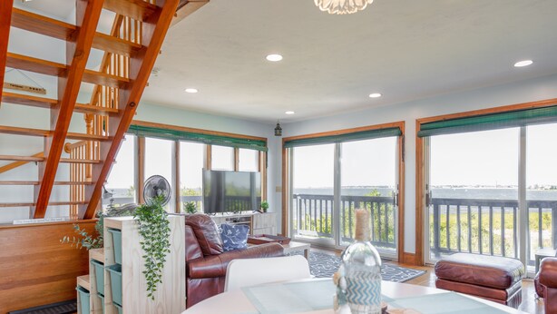 A spacious living area with a wooden staircase, a large window with a view, a TV, and a cozy seating arrangement. - Cozy Beach House with 270° Spectacular Views (Fairhaven)