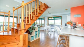 A spacious kitchen with a wooden staircase, white bar stools, and a large island with a sink. - Cozy Beach House with 270° Spectacular Views (Fairhaven)