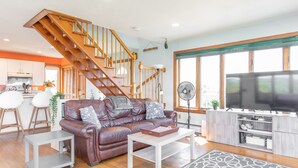 A spacious living room with a brown leather sofa, a white coffee table, a TV stand, and a wooden staircase. - Cozy Beach House with 270° Spectacular Views (Fairhaven)