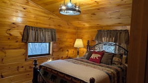 A wooden cabin bedroom with a bed, curtains, a lamp, and a chandelier.