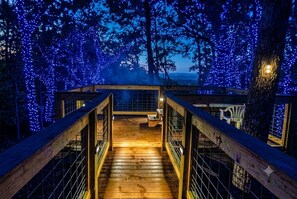 A wooden deck with railings leading to a covered area, surrounded by trees adorned with blue lights. - Enjoy the Holidays! Beautiful View! Hot Tub,Pet Friendly,9.8🌟










 (Sevierville)