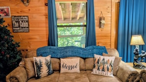 A cozy cabin interior with a wooden wall, a window with curtains, a couch with forest-themed pillows, and a small table with a lamp. - HAPPY FALL! Fall Special! Beautiful View! Hot Tub,Pet Friendly,9.8🌟










 (Sevierville)