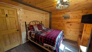 Une pièce aux murs lambrissés, comprenant un lit, une porte en bois, un luminaire au plafond et un téléviseur.