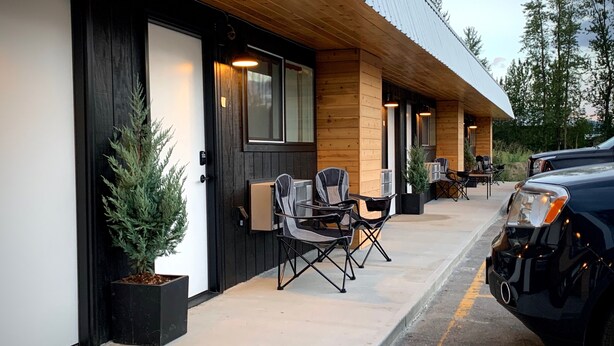 Un edificio con fachada de madera, un área de estar al aire libre techada con sillas, y un vehículo estacionado.