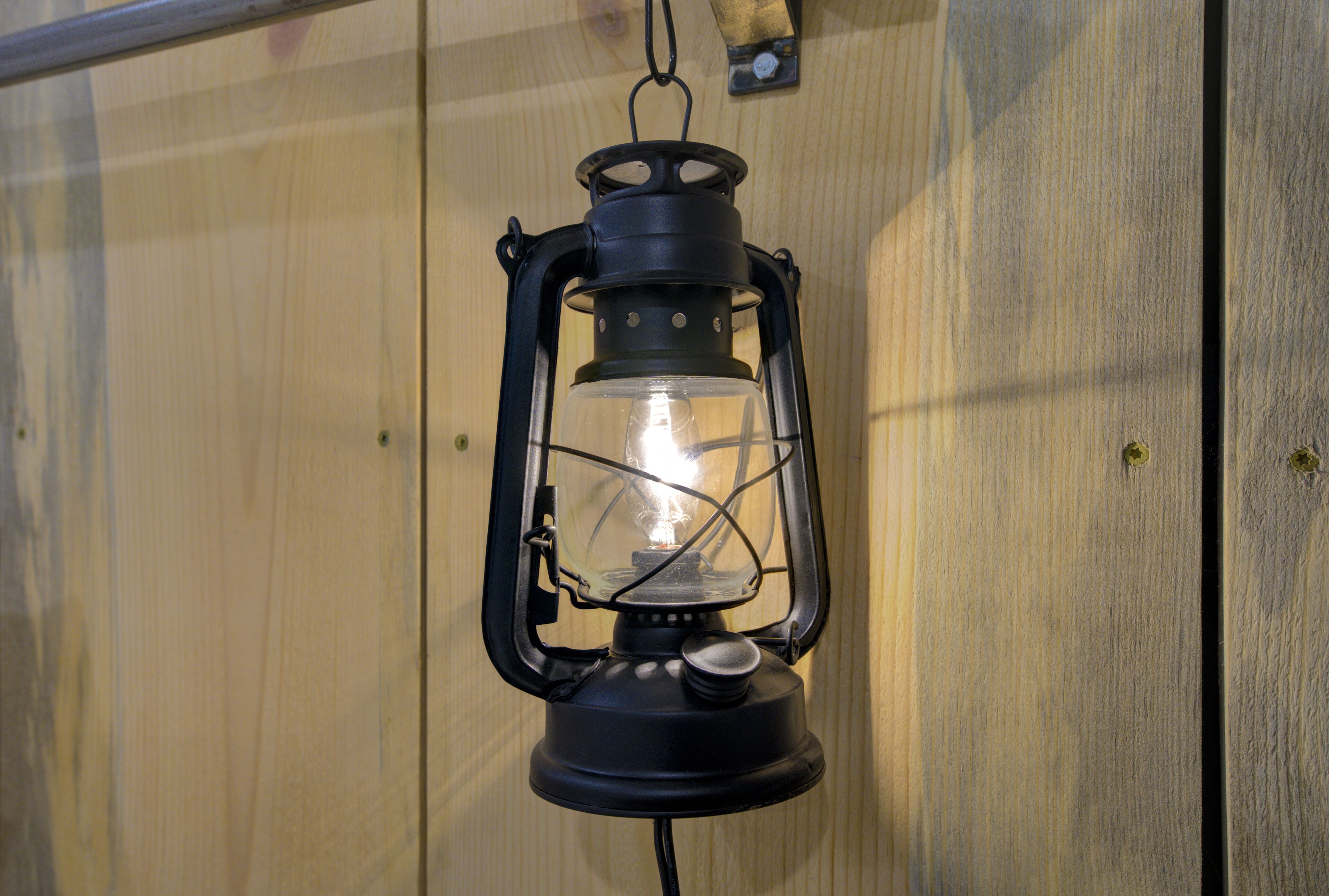 A black lantern with a clear glass enclosure and a visible light bulb inside, hanging against a wooden wall.