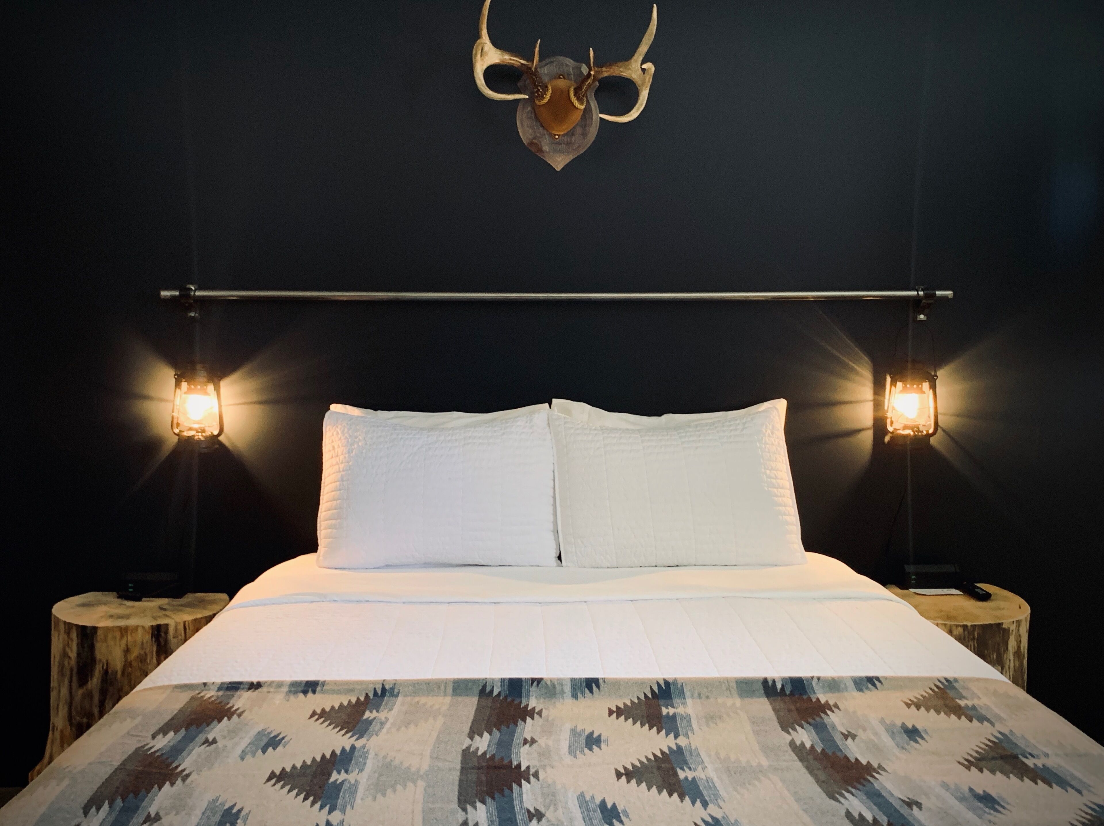 A bed with white linens, two bedside lamps, a wooden headboard, and a wall-mounted deer skull.