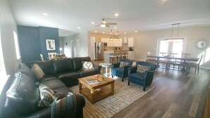 Ein geräumiges Wohnzimmer mit einem dunkelbraunen Ledersofa, einem Holztisch und einem blauen Sessel. Im Hintergrund ist eine Küche mit Essbereich zu sehen.