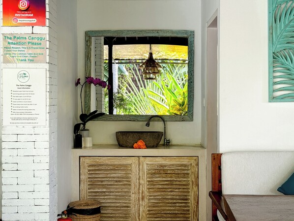 A bathroom with a rustic sink, a window with a view of greenery, and a sign with guest information. - The Palms Canggu (Canggu)